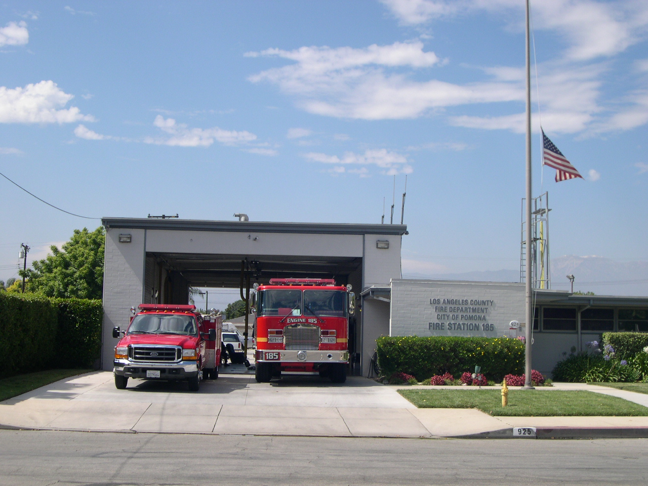 Bright red firetruck 185 next to regular truck carrying fire supplies. Both parked a bit outside of the station. Surrounded by clear skies and a beautiful day.